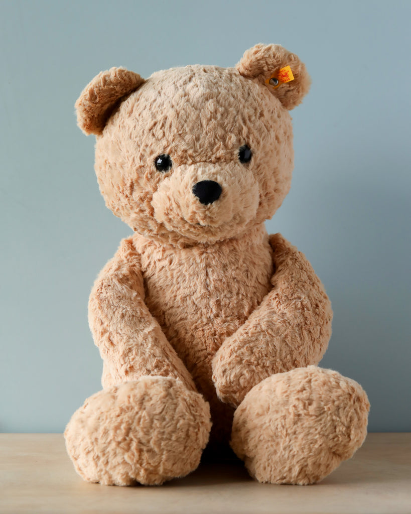 A Steiff XL Jimmy Teddy Bear with light brown fur is sitting against a pale blue background. The bear has dark, shiny eyes, a small nose, and a faint smile. A yellow tag is visible on the bear.