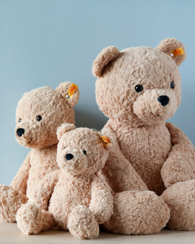 Three Steiff Jimmy Teddy Bears, 16 inches tall, in shades of tan, sitting together against a light blue background. The largest bear is at the back and the smallest in front. Each has a visible Button.
