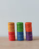 Three stacks of colorful Grapat 36 Coins in 6 Colors wooden coins in red, green, and blue hues, each decreasing in size from bottom to top, on a light wooden table against a grey background.