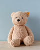 A Steiff Jimmy Teddy Bear, 16 Inches with a button in its ear sits on a wooden surface against a plain light blue background. The bear has a soft, textured coat and a friendly expression.