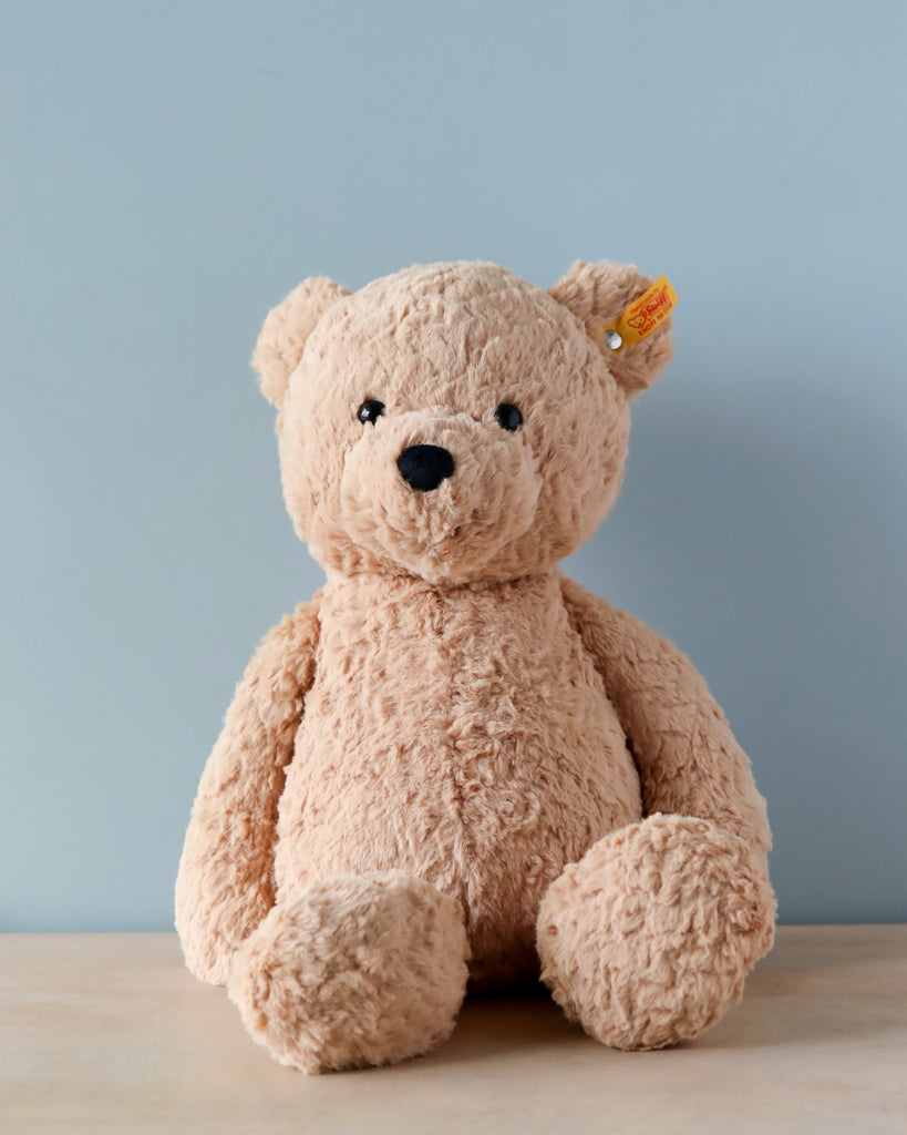 A Steiff Jimmy Teddy Bear, 16 Inches with a button in its ear sits on a wooden surface against a plain light blue background. The bear has a soft, textured coat and a friendly expression.