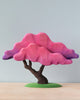 A colorful sculpture of a stylized, handmade Japanese Maple tree with a trunk in brown and wide, layered canopy in shades of pink and purple, handcrafted from linden wood, set against a simple blue