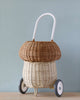 A hand-woven Olli Ella Rattan Mushroom Luggy - Natural with a white handle, resembling a small, quaint cart, displayed against a plain blue background.