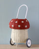 A whimsical Olli Ella Rattan Mushroom Luggy on wheels with a red top featuring white polka dots and a natural beige bottom, set against a soft blue background.