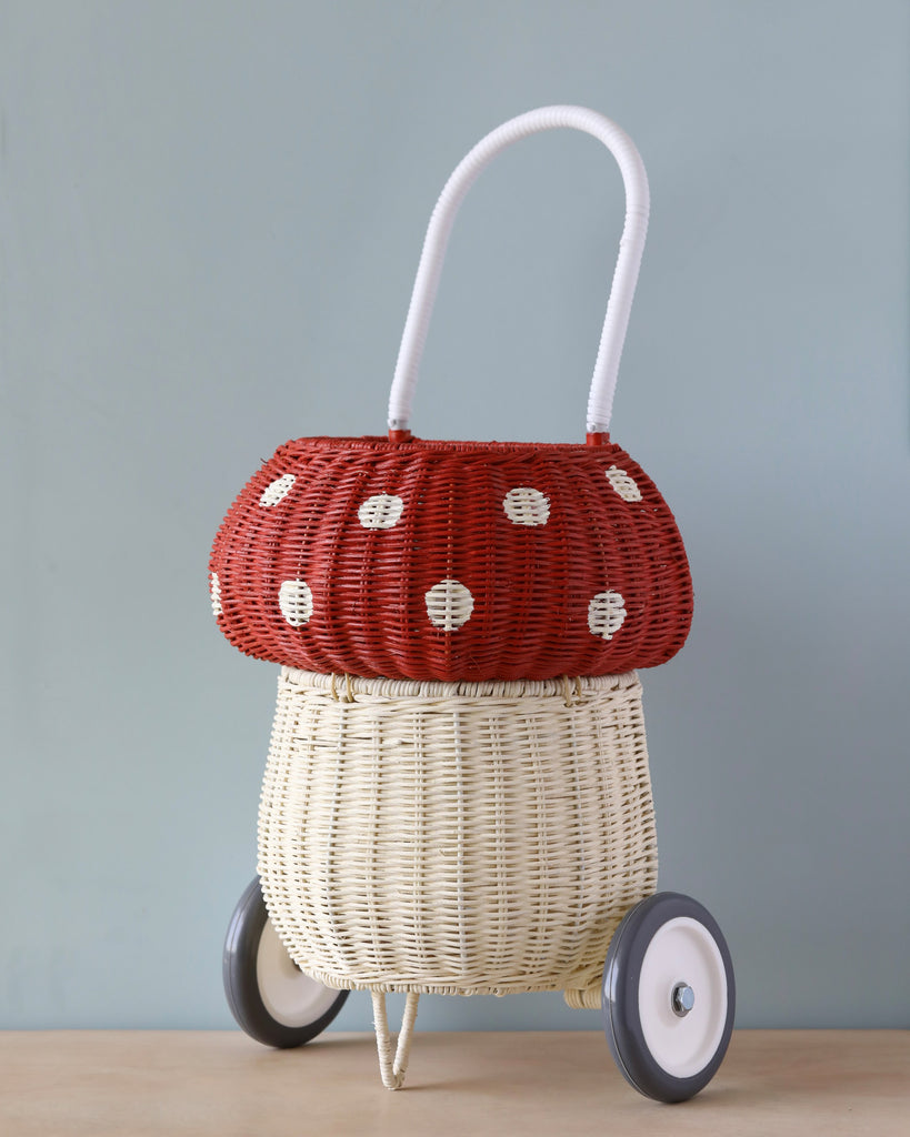 A whimsical Olli Ella Rattan Mushroom Luggy on wheels with a red top featuring white polka dots and a natural beige bottom, set against a soft blue background.