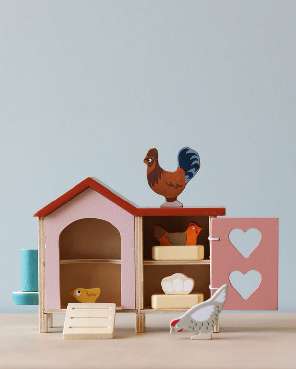 A small Chicken Coop with a roof, nesting boxes, and a door adorned with two heart-shaped cutouts. Wooden figures of a rooster and hens, along with chicks and freshly laid eggs, are arranged around and inside the Chicken Coop. One hen figure stands outside, pecking at the ground.