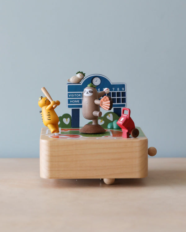 A colorful Baseball Music Box toy set depicting a basketball game. Features include a basketball hoop, scoreboard, a brown egg with limbs holding a ball, and other playful figures on a wooden base.