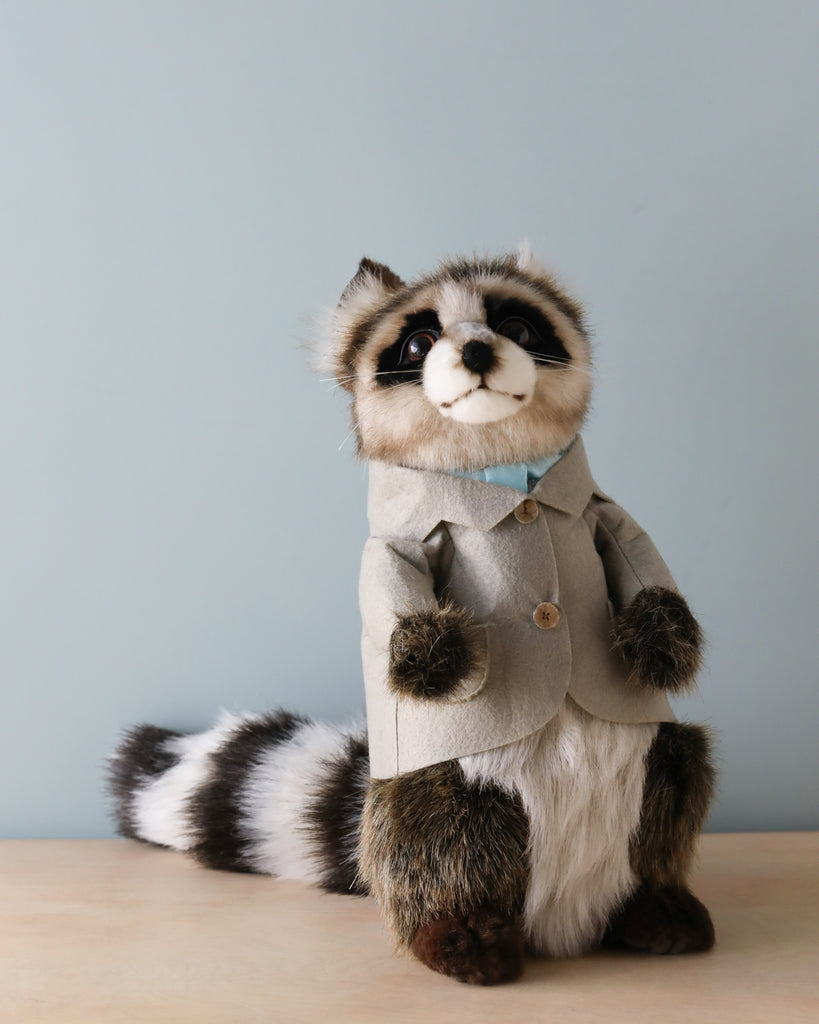 A Papa Raccoon With Jacket Stuffed Animal, hand sewn and dressed in a stylish gray coat, sitting upright against a pale blue background.