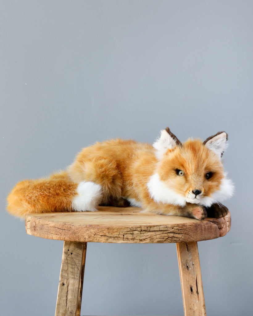 A Fox Stuffed Animal with artisan hand-sewn features lying on an old wooden stool against a plain gray background, with its head turned slightly to the side.