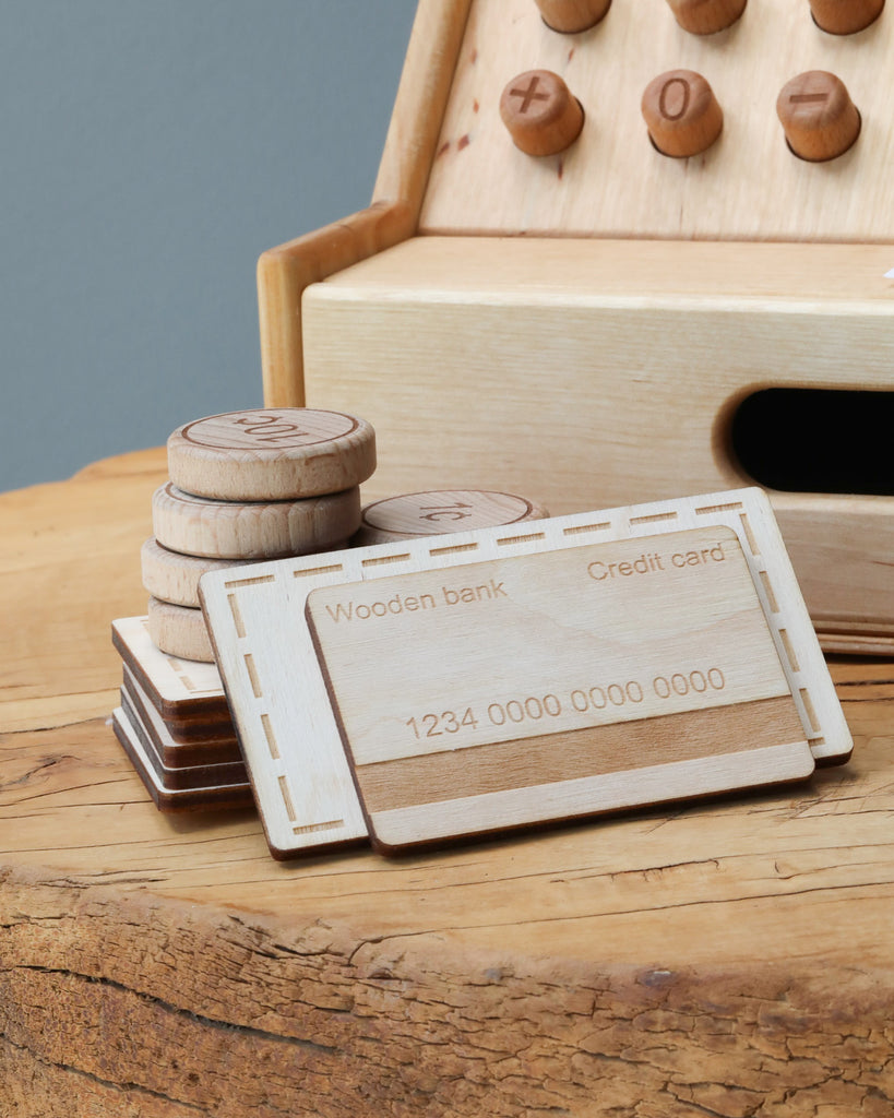 A wooden toy credit card and coins on a table, with a Wooden Cash Register in the background, simulating a finance or banking scenario for educational play.