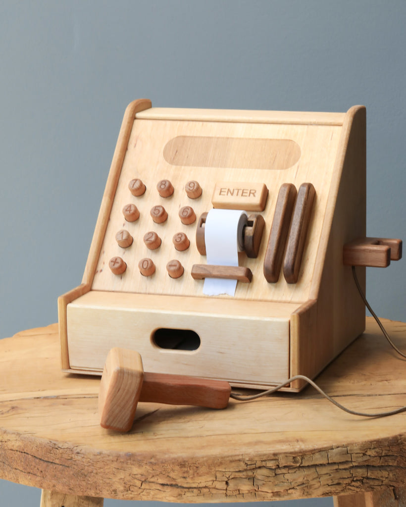 Wooden Cash Register children's toy featuring round buttons, a paper roll slot, and a lever, placed on a wooden table against a grey background.