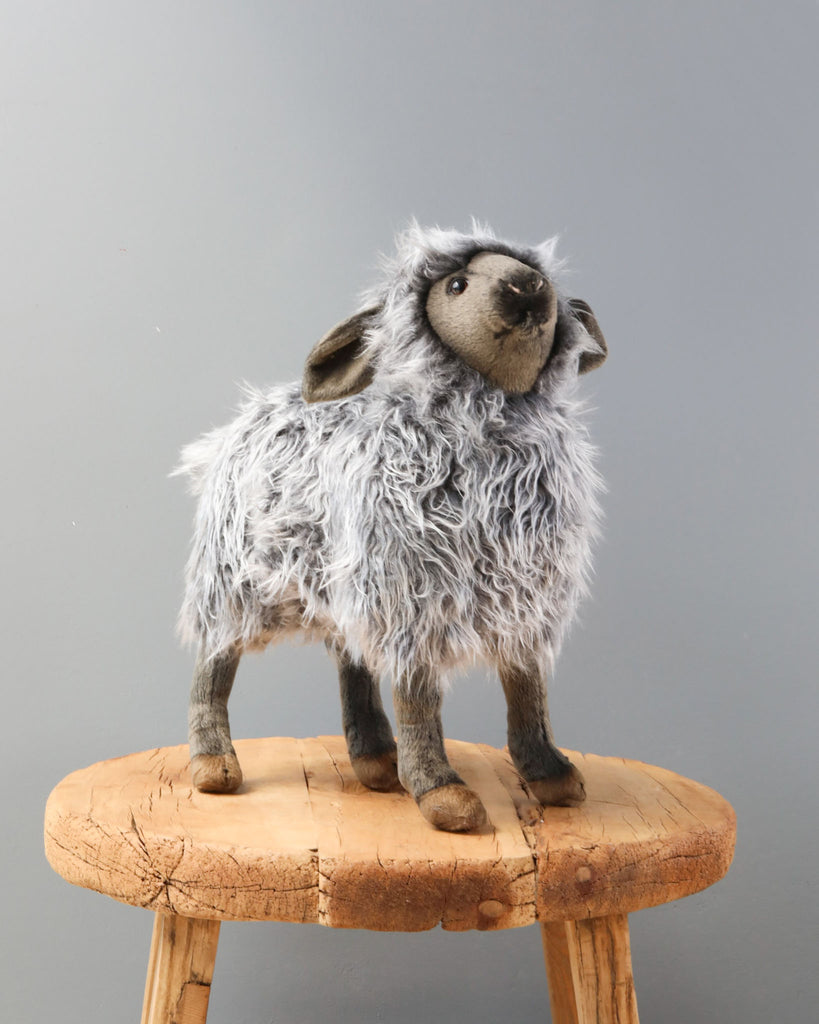 A hand-sewn Sheep Stuffed Animal with a fluffy gray body and darker gray face and legs, standing upright on a wooden stool against a neutral gray background.