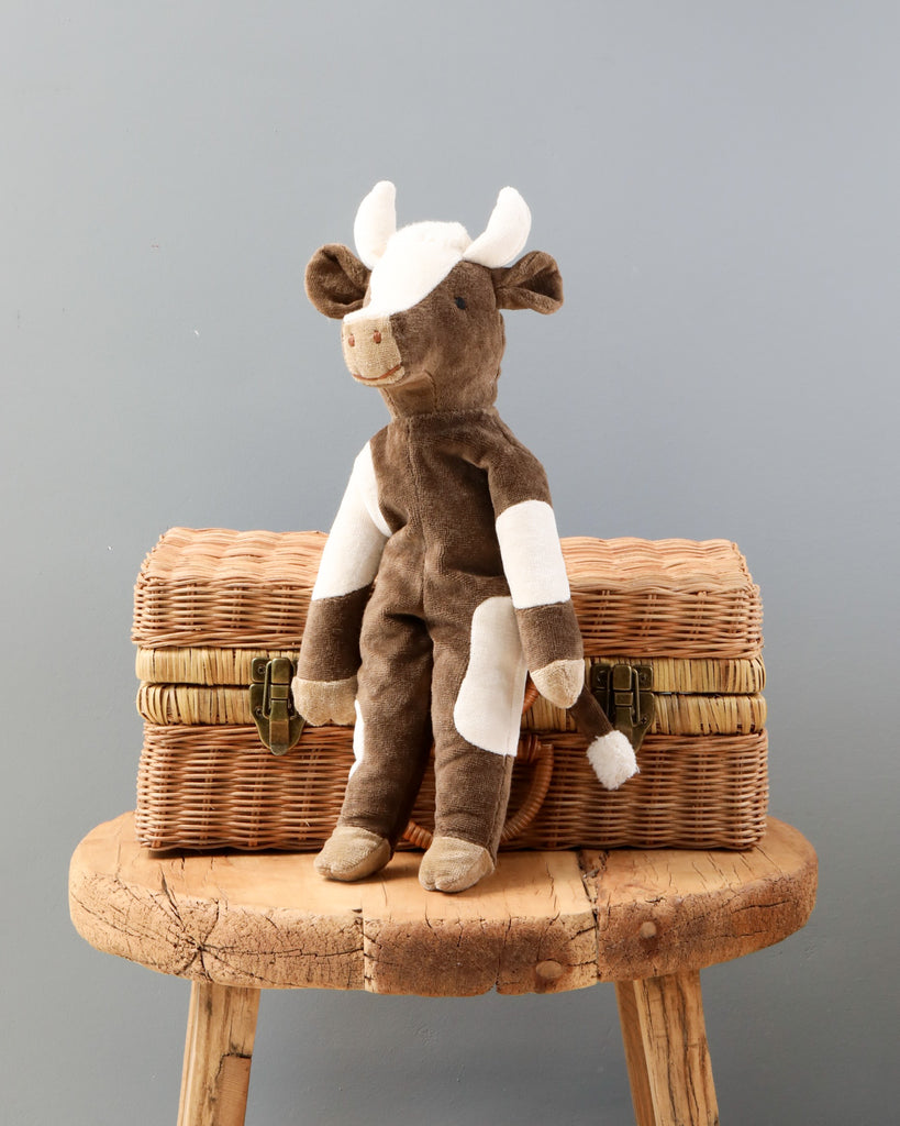 A Senger Naturwelt Stuffed Animal - Cow with white and brown patches sitting on a round, woven wicker basket placed on a wooden stool against a gray background.