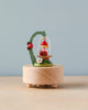 A charming, colorful hand cranked Mini Fairy On Swing music box featuring a small gnome sitting under an arched branch with a mushroom, adorned with a bird and flowers, placed on a wooden base against a soft blue background