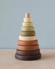 A Wooden Pyramid Stacker - Olive with rings in gradient shades ranging from dark brown at the base to light cream at the top, coated in non-toxic paint, set against a soft blue background.