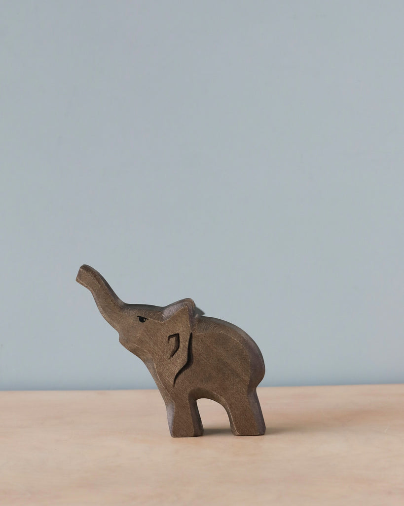 A Handmade Holzwald Baby Elephant wooden figurine stands on a tabletop, featuring intricate carvings and a lifted trunk, set against a neutral beige backdrop.