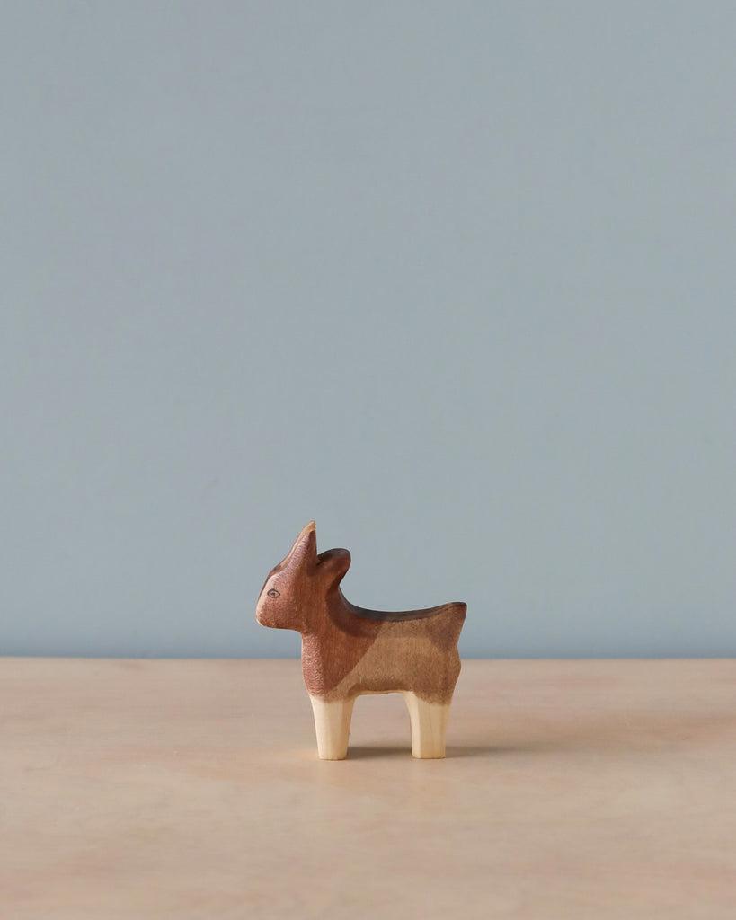 A small, high-quality wooden figurine of a Handmade Holzwald Small Goat standing against a plain blue backdrop with a light beige surface beneath.