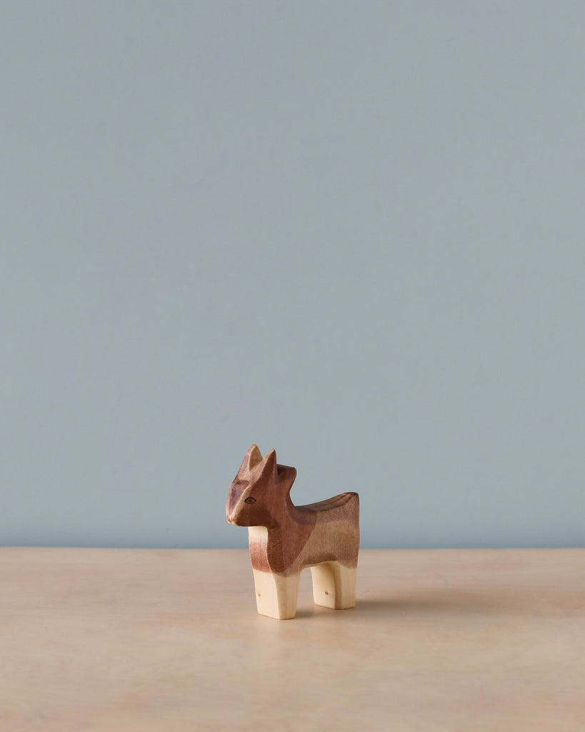 A small handmade Holzwald goat figurine, perfect for imaginative play, stands on a solid surface against a plain, light blue background. The goat is in a standing position, featuring distinctive grain patterns.