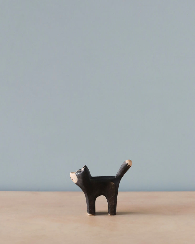 Sentence with product name: A minimalist photo featuring a small, Handmade Holzwald Black Cat against a light blue background. The cat has simplified, geometrical shapes and a visible wood grain pattern.