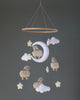 A handmade mobile featuring plush sheep, clouds, and a moon, all in soft white and cream colors, hanging from a wooden ring against a grey background.