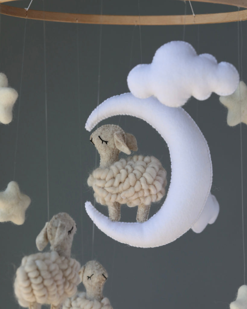 A soft, plush Handmade Mobile - Little Sheep - Final Sale nursery mobile with a sheep resting in a crescent moon amidst fluffy clouds against a gray background.