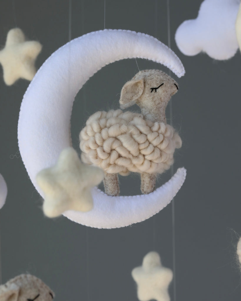 A felted Handmade Mobile featuring a fluffy sheep nestled inside a white crescent moon, surrounded by hanging felt stars, against a soft gray background.