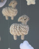 Close-up of a Handmade Mobile - Little Sheep - Final Sale ornament hanging by a thread from a ceiling mobile, with more blurred sheep figures in the background, depicting a peaceful and whimsical craft design.