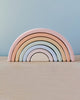 A set of Handmade Rainbow Stackers in pastel colors, designed for open-ended play, arranged in decreasing sizes to form a complete semicircle, displayed against a light blue and beige background.