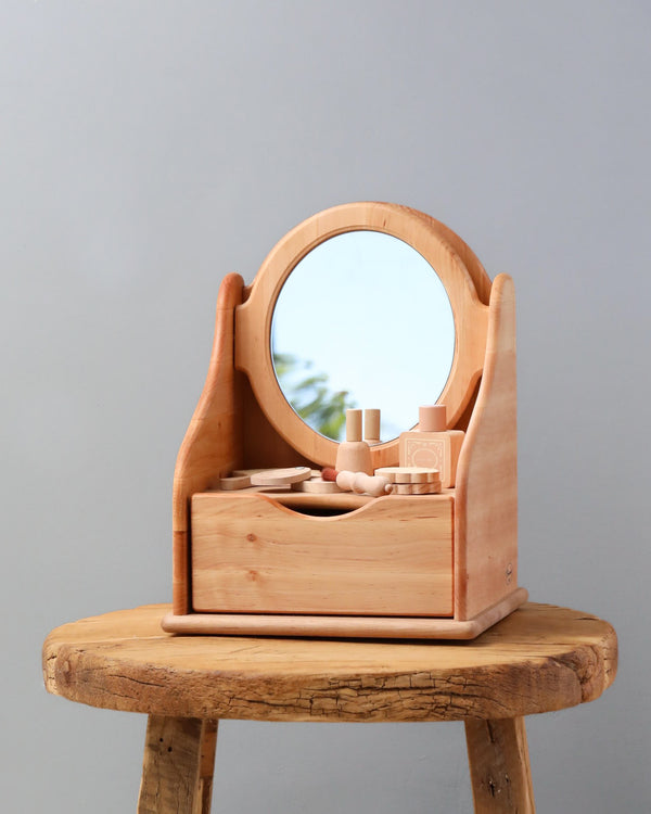 A Handmade Wooden Toy Vanity Table with a mirror, various compartments, and small beauty products, set on a round wooden stool against a plain gray background.