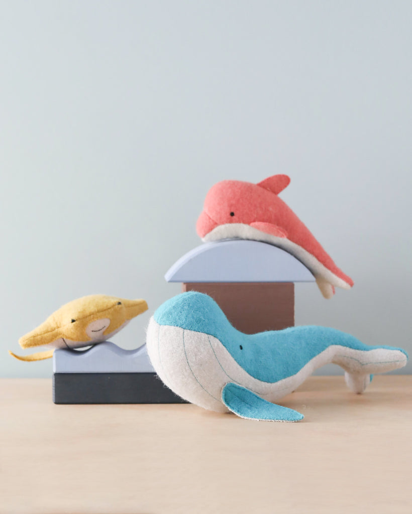 Three Olli Ella Holdie Folk Felt Ocean Animals - a blue whale, a red dolphin, and a yellow turtle - rest on a stack of books on a wooden table against a plain background. These handmade Ocean friends are perfect for imaginative.