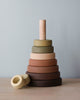 A Wooden Pyramid Stacker - Olive with rings in various earth tones neatly arranged on a spindle, one ring lying beside it on a light blue background, all coated in non-toxic paint.