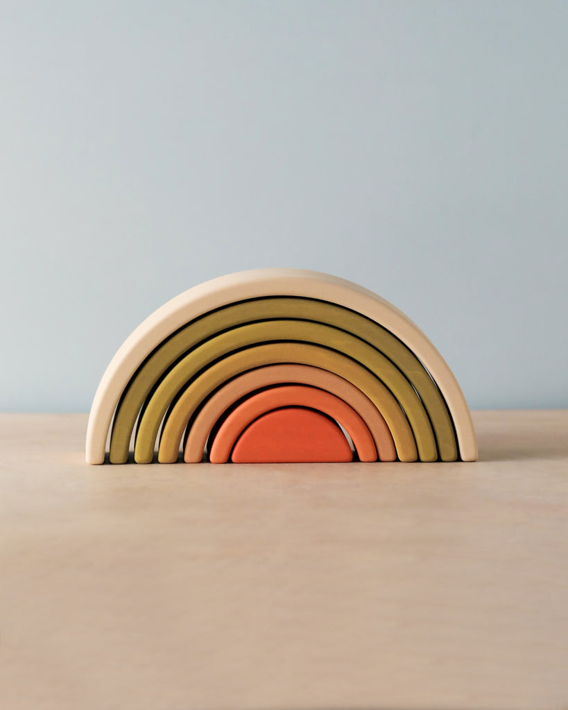 A Handmade Mini Rainbow Stacker - Flower Meadow with colorful arches arranged from largest to smallest on a wooden surface against a pale blue background.