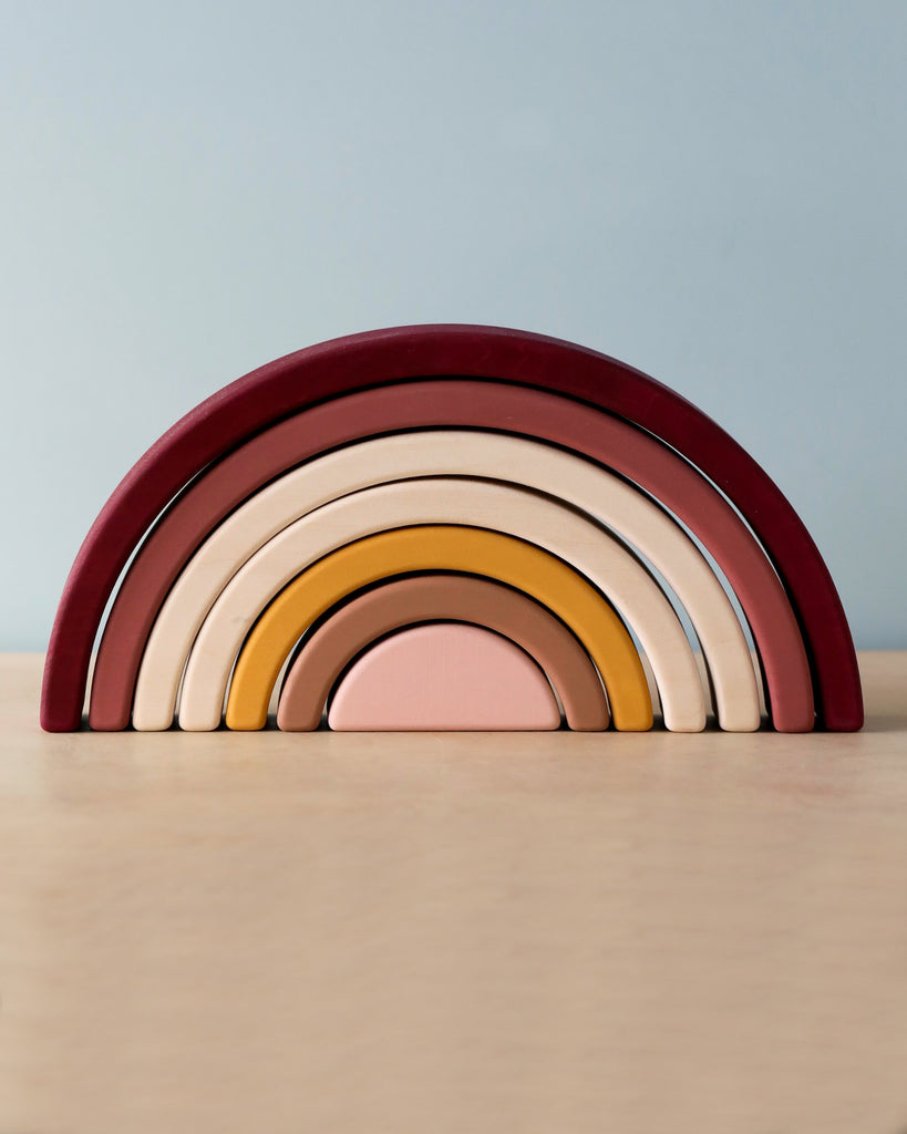 The Handmade Rainbow Stacker - Pink is a handmade stacking toy featuring arches in varying sizes and colors, arranged to form a rainbow shape. The colors range from muted reds and pinks to beige and mustard yellow. Crafted with non-toxic paint, the toy sits on a light-colored wooden surface against a pale blue background, perfect for open-ended play.