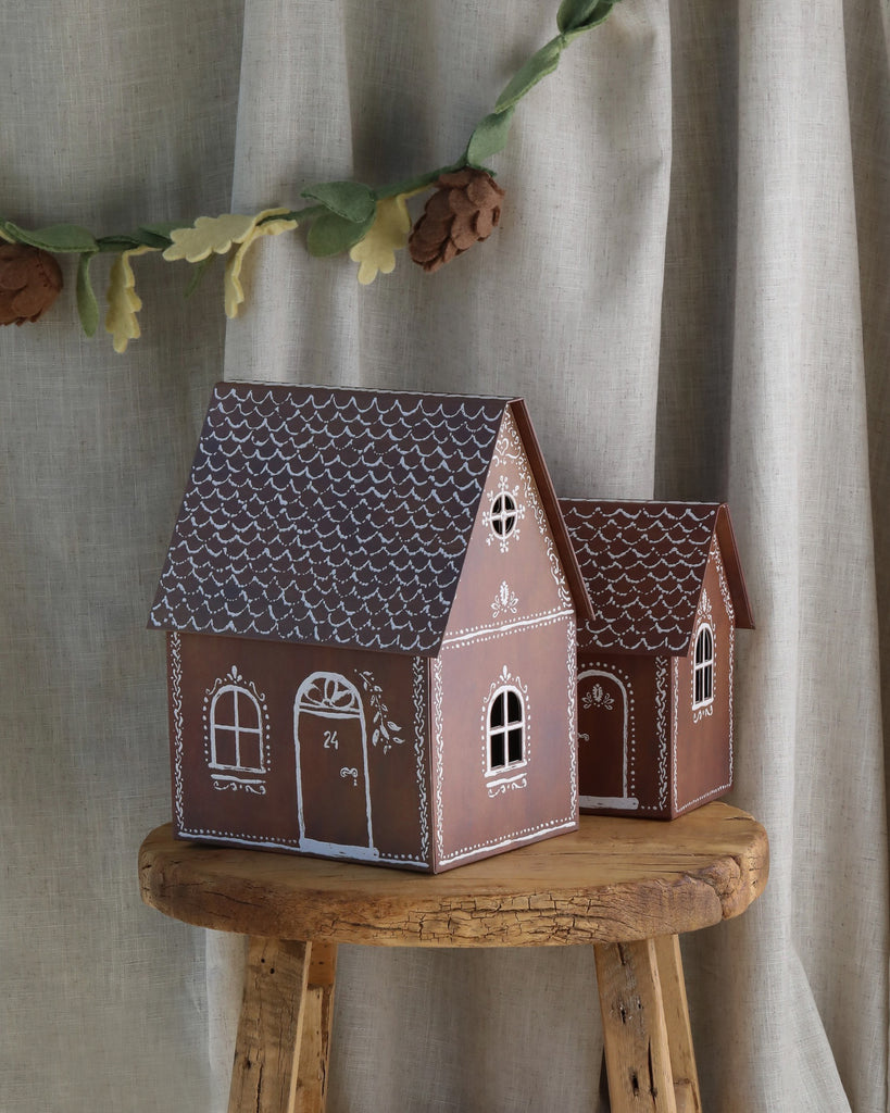 A decorative display features two intricately designed Maileg Cardboard Gingerbread Houses with white icing detailing. They sit on a wooden stool in front of a beige curtain, with a felt garland of autumn leaves and pinecones draped above, creating holiday surprises for your micro Maileg friends.