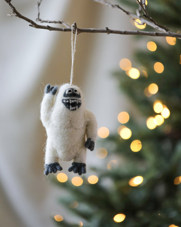 A Handmade Felt Yeti Christmas Tree Ornament hanging on a tree branch, with softly blurred Christmas lights in the background.