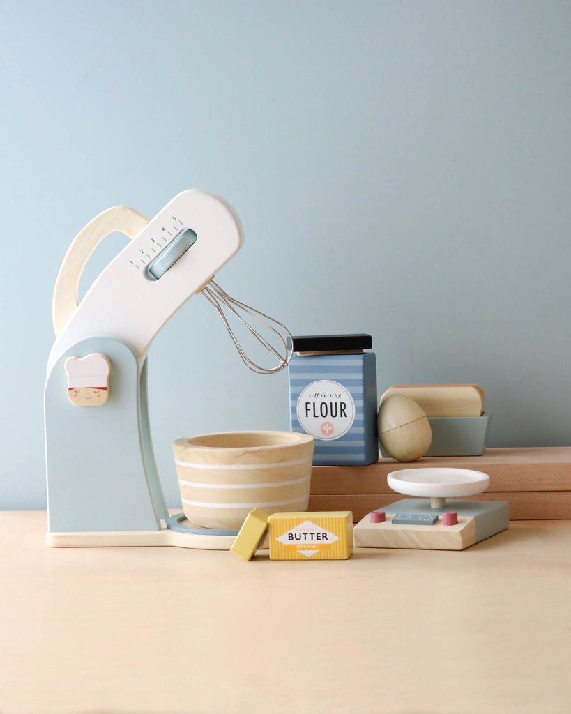 A stylish mixer in light blue on a kitchen counter with accessories including wooden bowls, a wooden spoon, and pretend food items like flour, butter, and eggs.