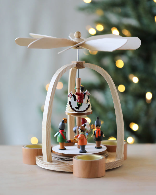 A traditional wooden Christian Ulbricht Two Tier Christmas Pyramid, handcrafted in Germany, featuring small figurines of a horse and children, set under a carved helicopter blade. The setup is on a wooden table with tea light candles.