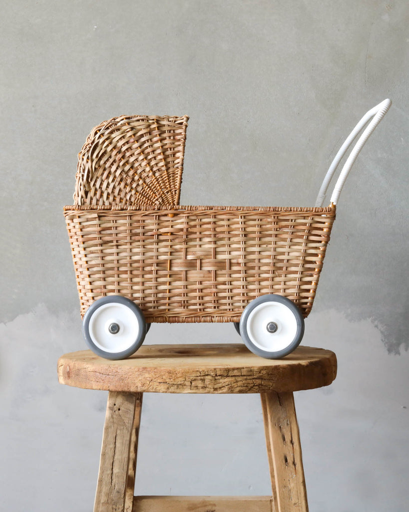 A hand-woven Olli Ella Rattan Doll Stroller with white wheels, resting on a rustic wooden stool against a textured gray wall. The carriage features a classic, handcrafted design.