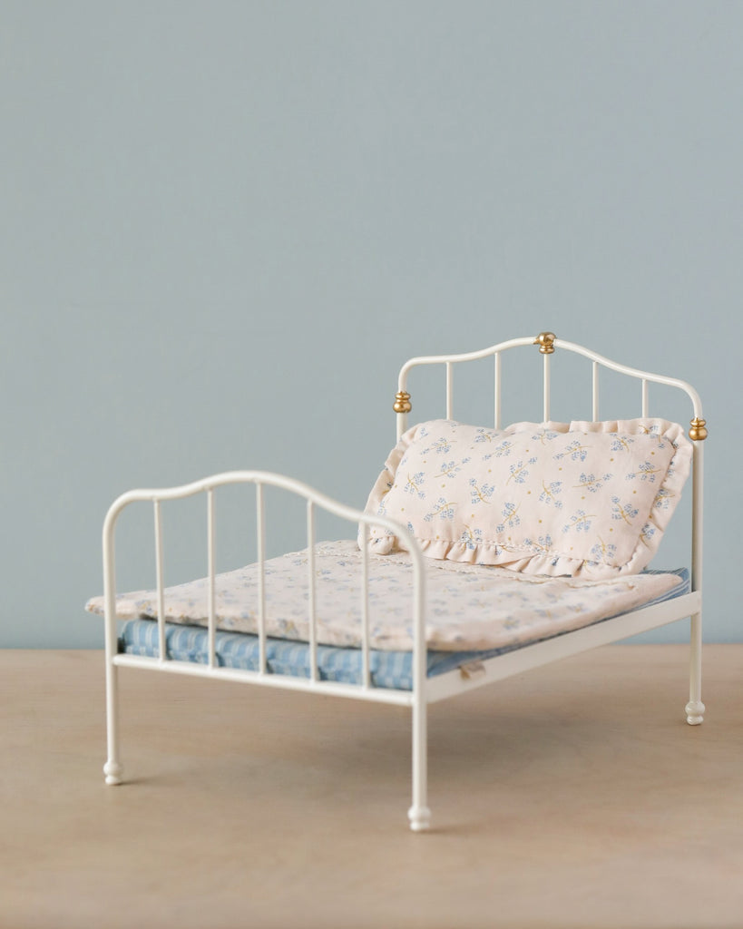 A Maileg Bed, Parent Mouse - Off White with straight lines and brass accents on the headboard and footboard. The bed, ideal as a bed for bigger mice, is dressed with a light blue mattress, a white comforter with Maileg printed fabrics featuring a delicate floral pattern, and a matching pillow, all set against a simple gray background.