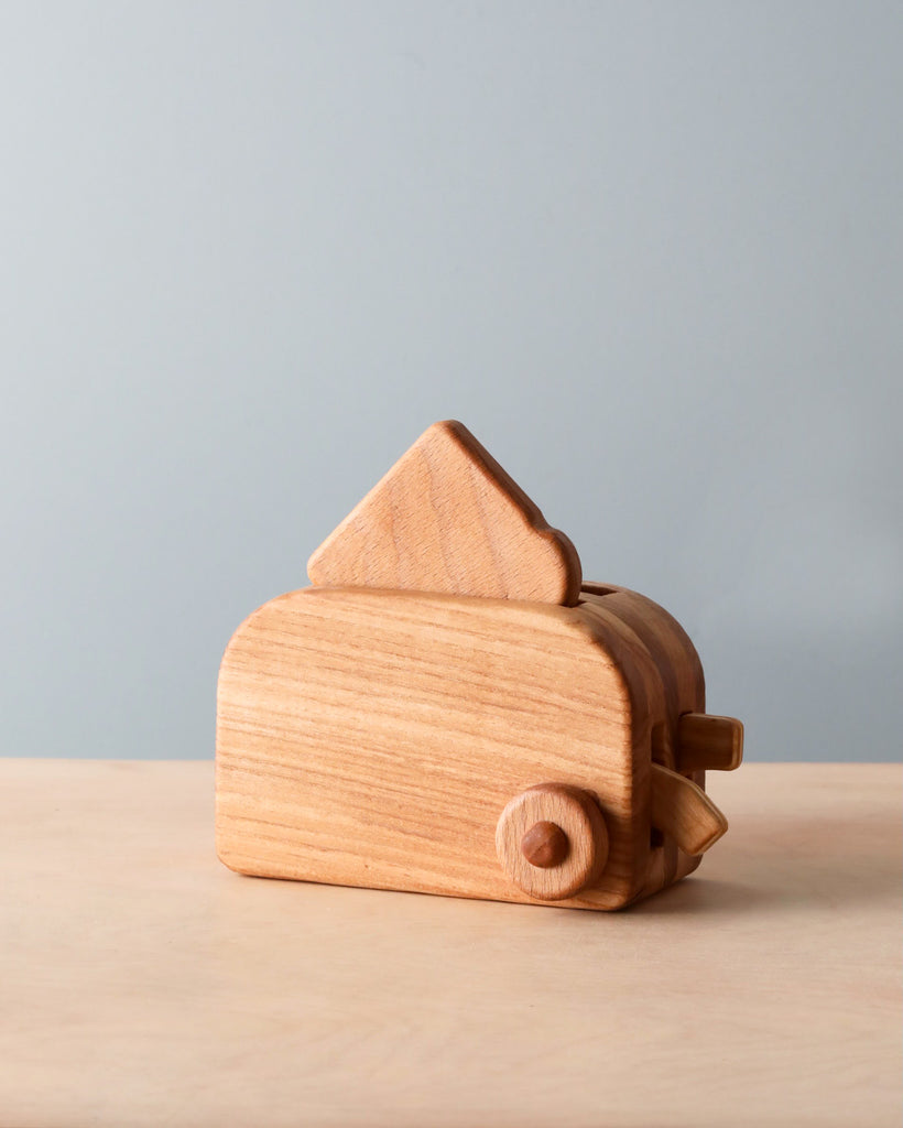 A Handmade Wooden Toaster shaped like a house, with a simplistic design and smooth finish, placed against a neutral gray and pale blue background.