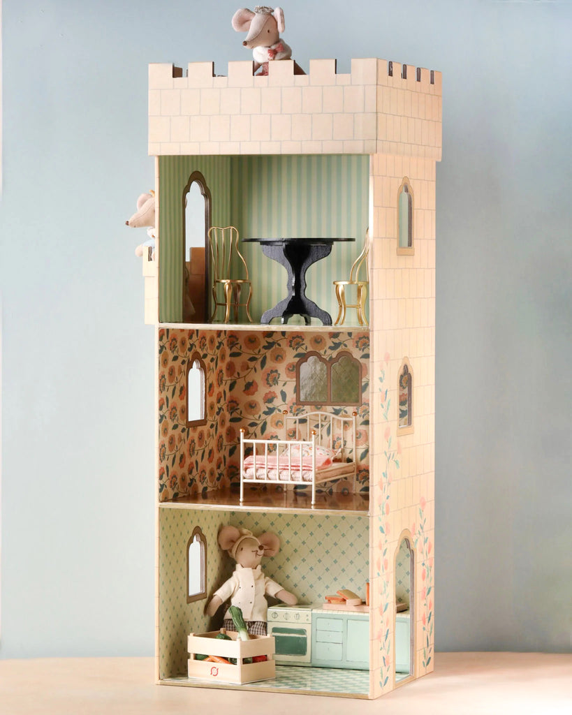 A three-story wooden Maileg Castle Tower styled as a castle for mice features Maileg prints. The house includes mice figurines in different rooms: an upper room with chairs and a table, a middle room with a crib, and a lower room serving as a kitchen with appliances and a mouse holding a basket.