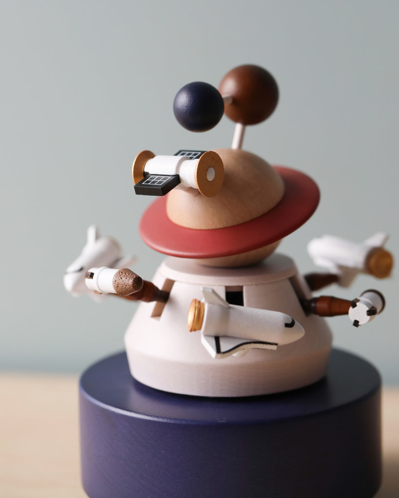 A whimsical Wooden Outer Space Music Box of a small figure with a spherical head and multiple arms, each holding different objects like a paintbrush and rolls of paper, placed on a circular stand that features an integrated hand