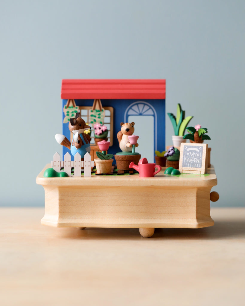A miniature display featuring two animal figurines, possibly beavers, tending to a gardening table filled with plants, pots, and gardening tools, set against a soft blue background. This charming scene is the Wooden Fox Gardener Music Box.