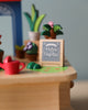 A close-up of a Wooden Fox Gardener Music Box sign that reads "#my garden" on a play table with small toy plants and a watering can in the background.