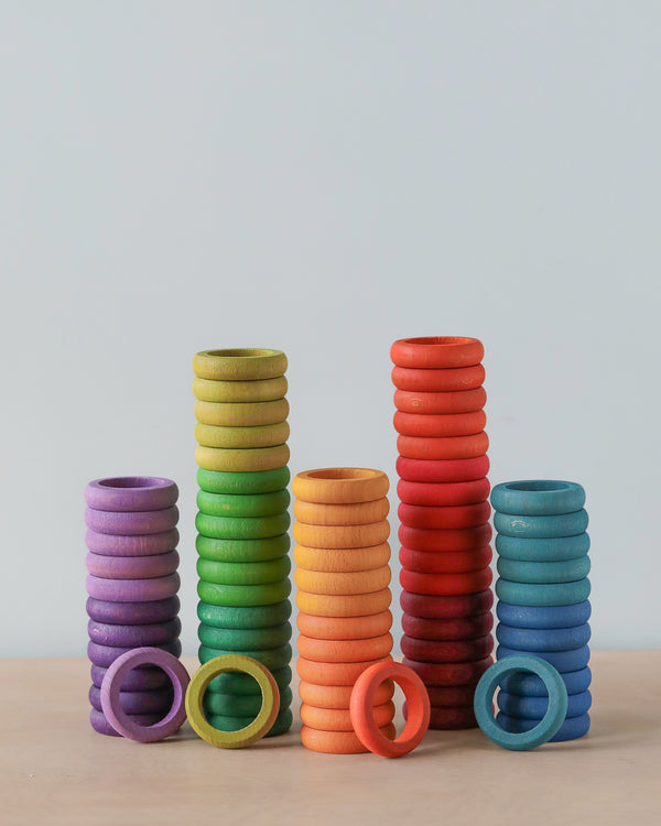 Five stacks of Grapat 72 Rings in 12 Colors arranged on a light wooden table against a neutral background, progressing in colors of purple, green, orange, red, and blue, ideal for sorting and imaginative.