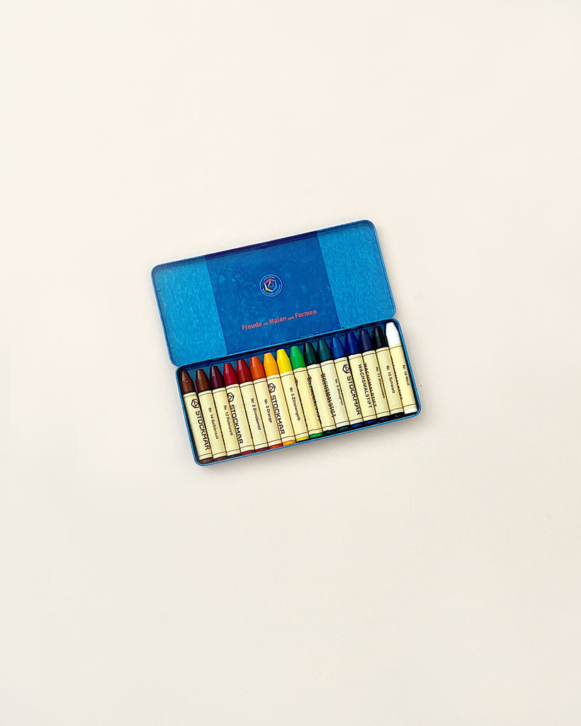 A set of Stockmar Wax Stick Crayons Tin Case - 16 Assorted neatly arranged in a blue, open metal case against a plain white background. Each crayon is labeled with a different color name.