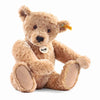 A handmade Steiff Elmar Teddy Bear with a fluffy golden-brown fur, sitting upright and displaying its iconic Button in Ear and a chest tag with the brand name.