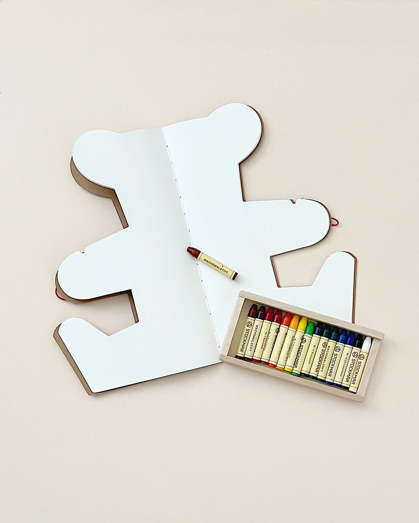 An open, puzzle-shaped Teddy Bear Drawing Book with crayons inside, displayed on a plain background. A red crayon is placed on top of the kit to decorate it.