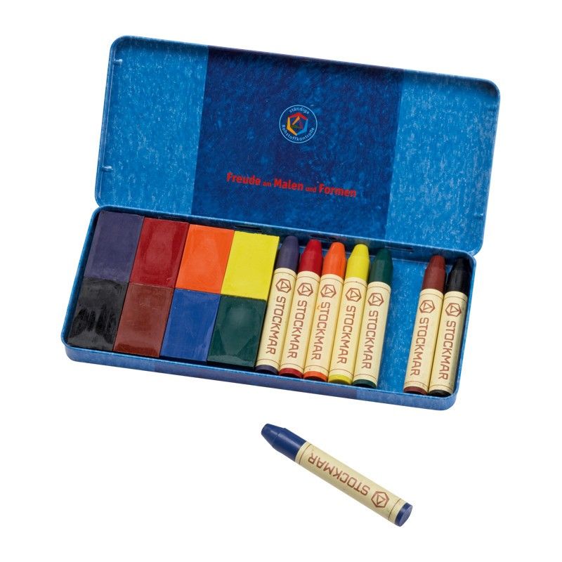 An open box of Stockmar Wax Crayons Combo Standard Tin Case with 8 Blocks & 8 Sticks Assorted neatly aligned inside and one block lying outside the box on a white surface. The lid and box are blue with a logo on the lid.