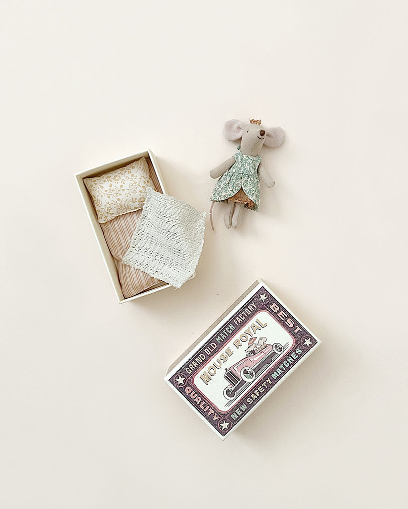 A Maileg Princess Little Sister Mouse with a floral-patterned skirt and a golden tiara, next to an open matchbox filled with miniature bedding, set against a light beige background.